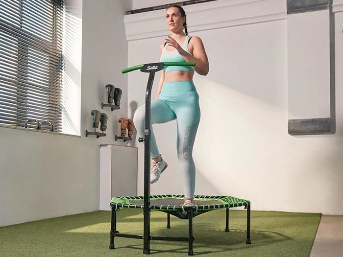 En kvinde i turkis træningstøj træner på en minitrampolin med styr i et veloplyst indendørs fitnesscenter.