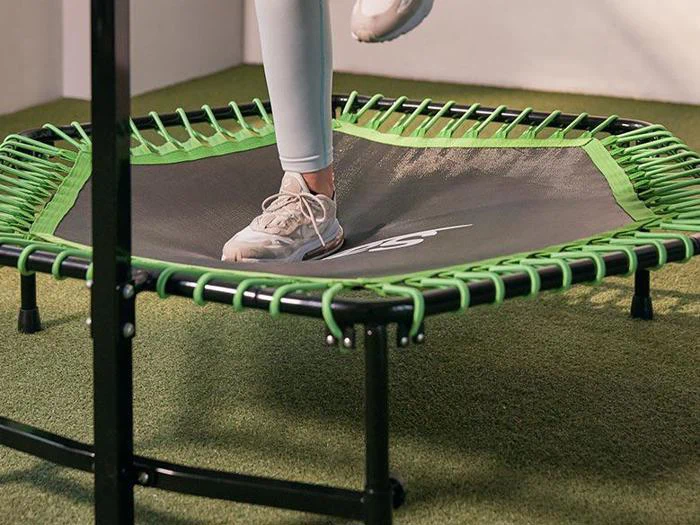En person i lyse leggings og sneakers træder på en lille sort og grøn trampolin indendørs på kunstgræs.