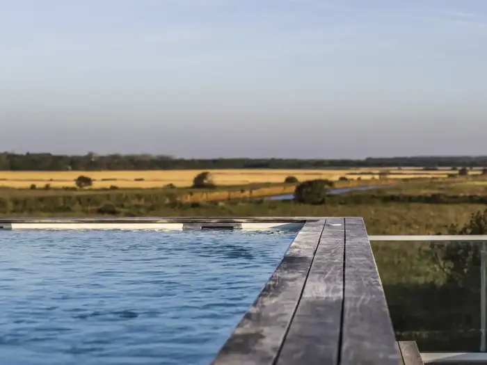 En udendørs pool med træterrasse har udsigt over marker og fjerne træer under en klar himmel.