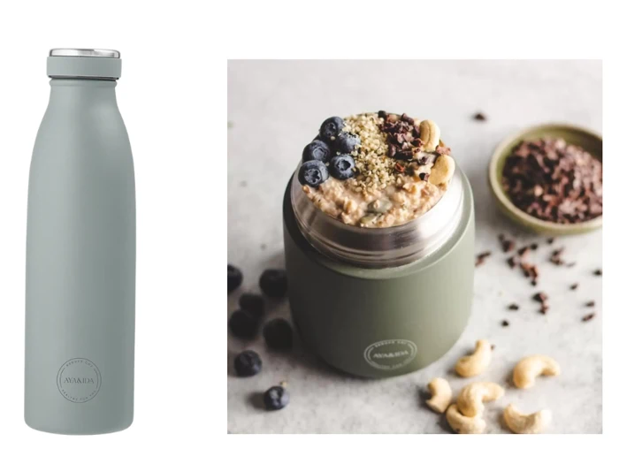 En salviegrøn isoleret flaske og en matchende madkrukke fyldt med havregryn, blåbær, cashewnødder og frø, med ekstra blåbær og en skål med kakaonibs i nærheden.