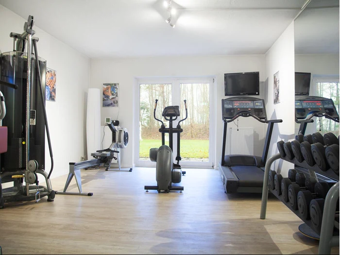 Et lyst træningscenter med træningsmaskiner, herunder løbebånd, crosstrainer, romaskine, vægtstativ og væghængt tv, der vender ud mod store glasdøre med udsigt til det fri.