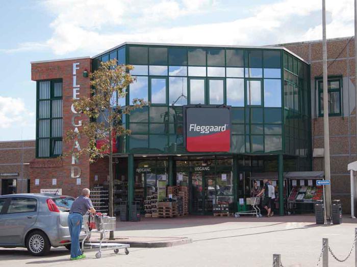 En person skubber en indkøbsvogn uden for Fleggaard, et moderne supermarked i mursten og glas, på en solrig dag.