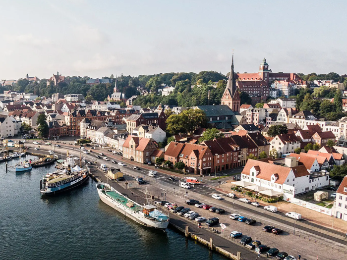 Luftfoto af en europæisk kystby med bygninger med røde tage, en kirke, parkerede biler og både, der ligger ved kajen.