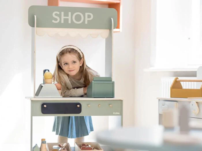 En ung pige står smilende bag disken i en legebutik med legetøjsvarer og et kasseapparat i et lyst rum.