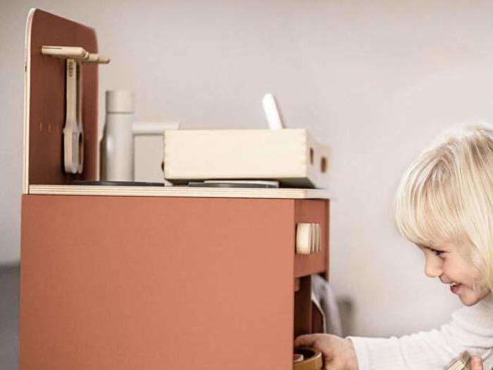 Et lille barn med blondt hår leger med et legetøjskøkken i træ, smiler og interagerer med legeapparaterne.