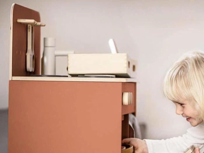 Et lille barn med blondt hår leger med et legetøjskøkken i træ, smiler og interagerer med legeapparaterne.