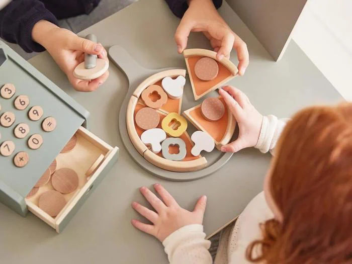 To børn leger med et pizzasæt af træ, hvor de samler skiver med forskelligt fyld, og ved siden af dem på et bord ses træmønter.