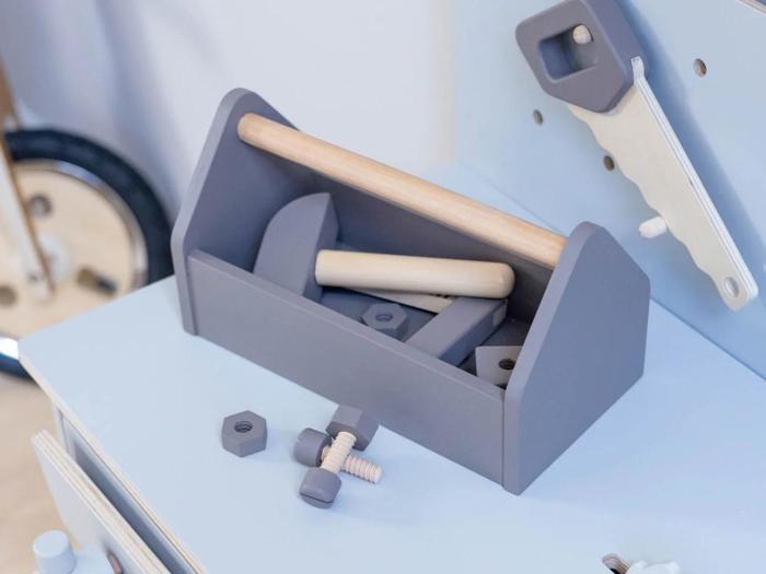 En grå legetøjsværktøjskasse af træ med håndtag, legetøjsværktøj og bolte står på en lyseblå flade med en legetøjssav og en del af et hjul i baggrunden.