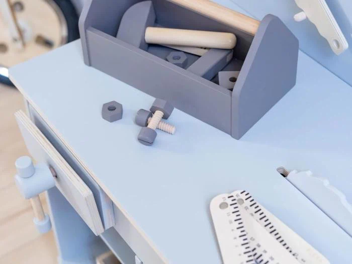 Et lyseblåt legetøjsarbejdsbord med en grå værktøjskasse og legetøjsskruer, -bolte og -nøgler af træ spredt på overfladen.