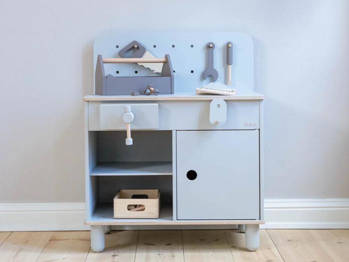 Et legearbejdsbord i træ til børn med legetøjsværktøj, en værktøjskasse og opbevaringshylder i lyseblå og grå, placeret mod en lys væg.