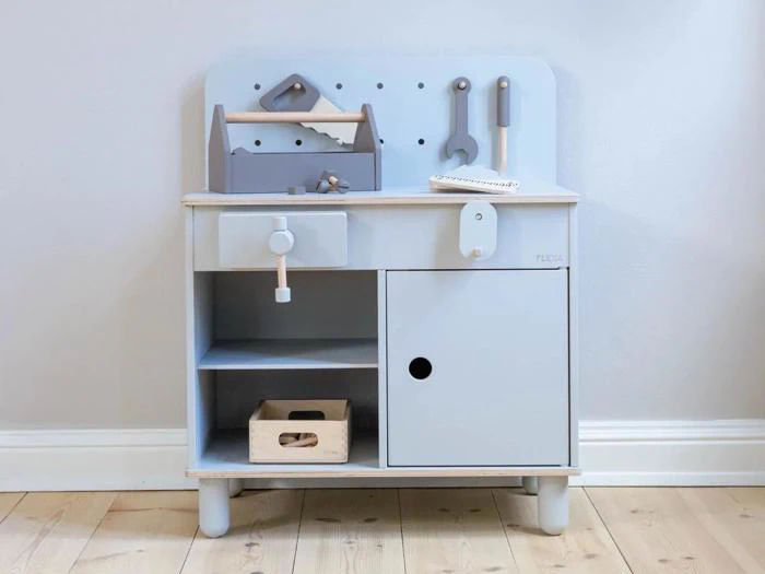Et lyseblåt legetøjsarbejdsbord af træ med værktøj, en værktøjskasse, hylder og et skab, opstillet mod en lys væg på et trægulv.