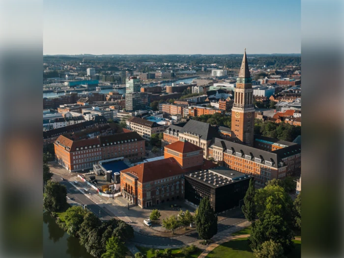 Luftfoto af et bybillede med røde murstensbygninger, et højt klokketårn og omkringliggende grønne områder under en klar himmel.