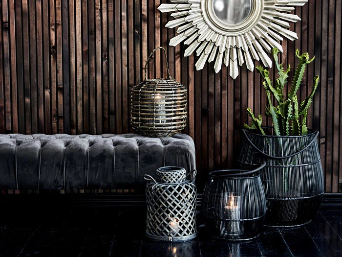 Et moderne interiør med en tuftet grå bænk, et spejl med solindfald, flettede lanterner og en høj kaktus i potte mod en mørk træpanelvæg.