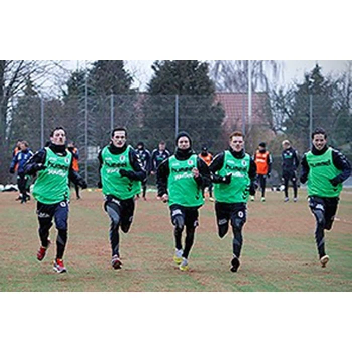 Fem fodboldspillere iført grønne træningstøj løber sammen på en græsbane under træning, med træer og andre spillere i baggrunden.