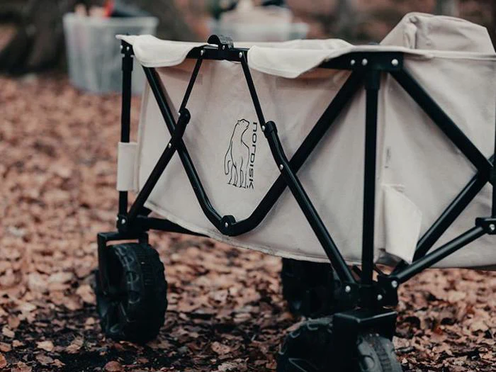 En beige sammenklappelig vogn med sort stel og robuste hjul står på nedfaldne blade. Vognen har et ulvelogo og ordet NORDISK på siden.