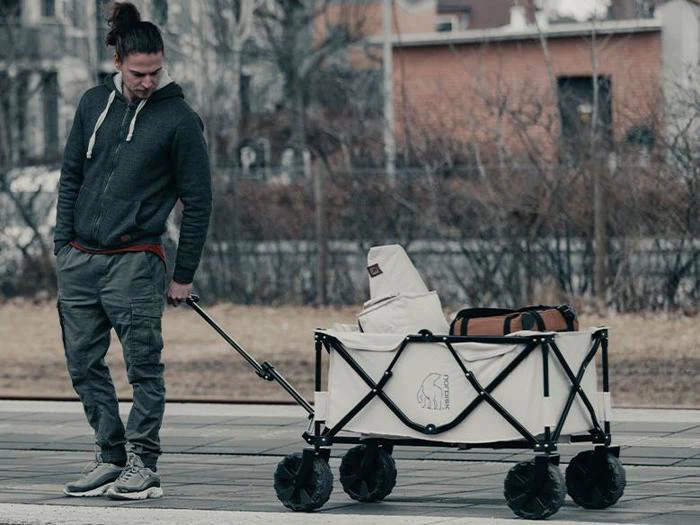 En mand i fritidstøj trækker en læsset klapvogn på et fortov; vognen indeholder tasker og friluftsudstyr.