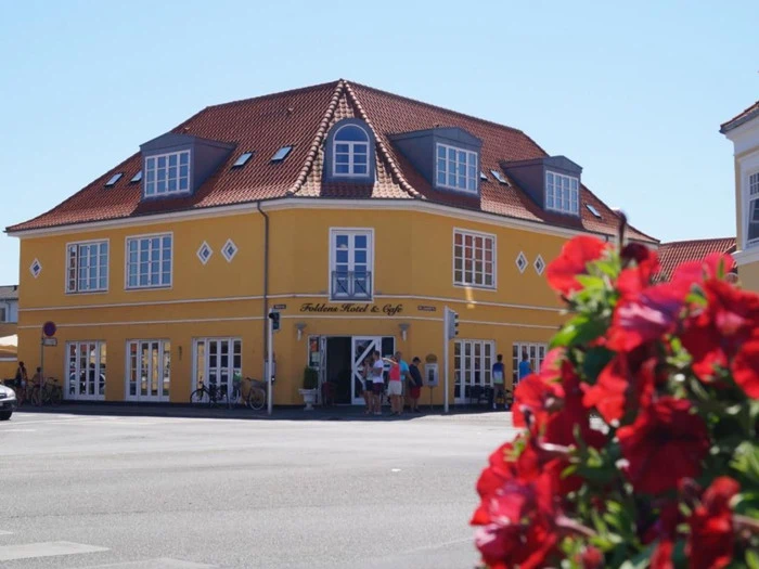 En gul toetagers hjørnebygning med kvistvinduer huser et hotel og en café. Røde blomster er slørede i forgrunden, og flere mennesker står nær indgangen.