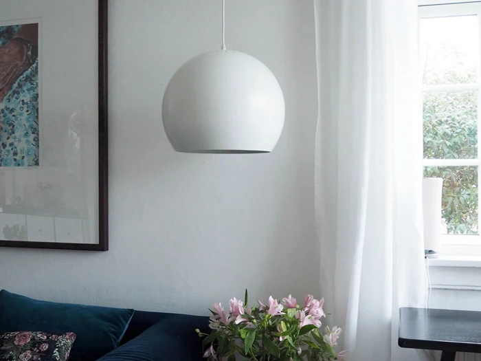 En hvid, kugleformet pendel hænger ned fra loftet over en sofa med en pude med blomstermønster og lyserøde blomster, ved siden af et vindue med tynde, hvide gardiner.