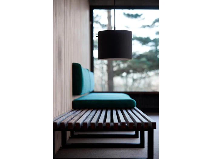 En moderne træbænk med grønne hynder sidder ved siden af en træbeklædt væg under en sort pendellampe med store vinduer, der viser træerne udenfor.