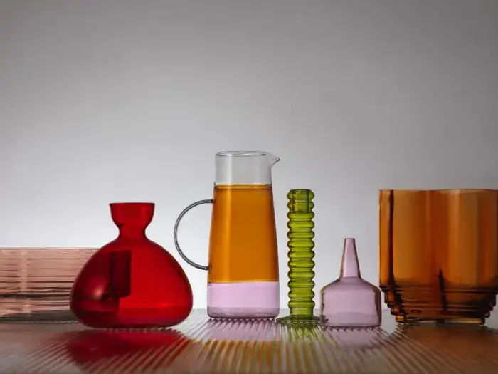 Forskellige farverige glaskar, bl.a. en rød vase, en klar kande med ravfarvet væske, en grøn ribbet cylinder, lyserøde flasker og et ravfarvet glas, arrangeret på en stribet overflade.