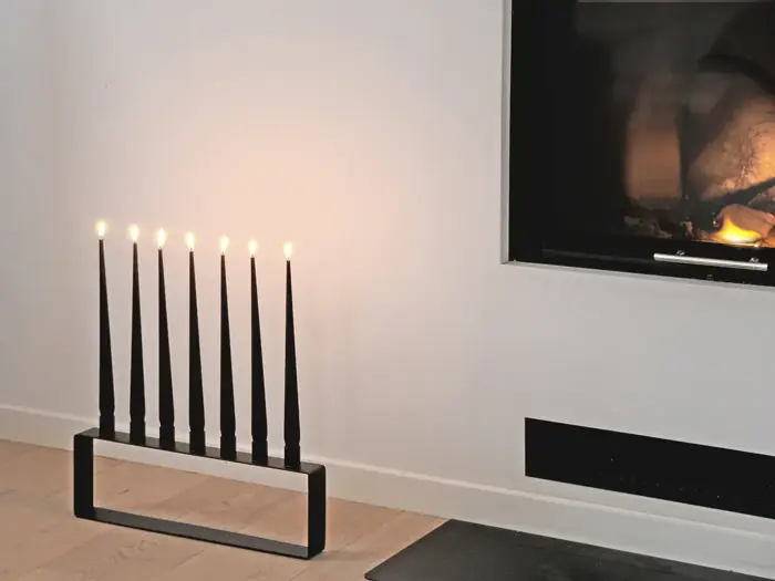 En moderne sort lysestage med seks tændte stearinlys står på et trægulv nær en pejs med en lille flamme indeni.