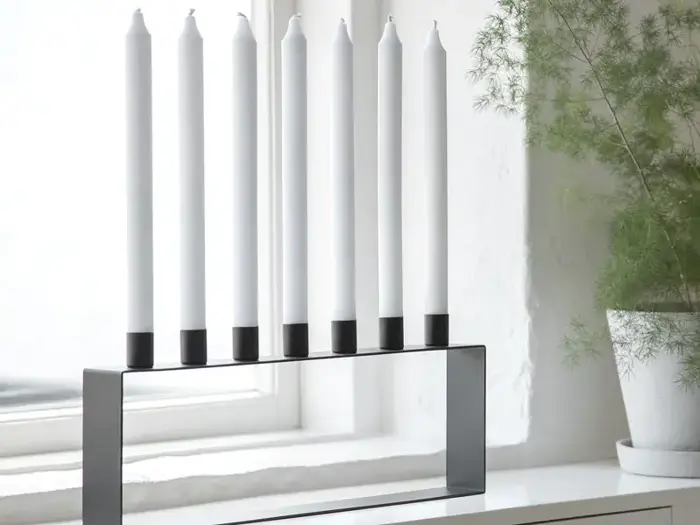 En moderne, minimalistisk menorah med syv utændte hvide lys står i vindueskarmen ved siden af en grøn potteplante.