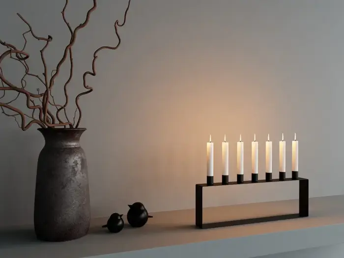En keramikvase med krøllede grene, to små sorte fuglefigurer og en moderne sort lysestage med seks tændte lys på en grå flade mod en ensfarvet væg.