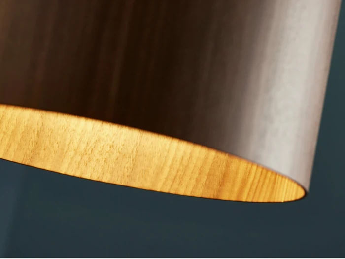 Nærbillede af den nederste kant af en cylindrisk lampeskærm med træstruktur, blødt belyst indefra, mod en mørk, sløret baggrund.