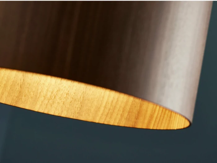 Nærbillede af en moderne, cylindrisk lampeskærm af træ med varmt lys, der stråler indefra mod en mørk baggrund.