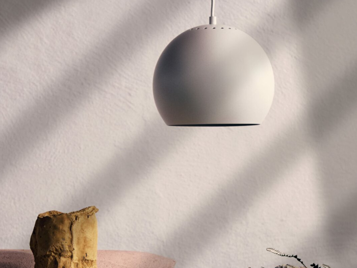 En moderne, kugleformet hvid pendel hænger fra loftet og kaster bløde skygger på en struktureret hvid væg. En lille sten og en plante er synlige nedenunder.