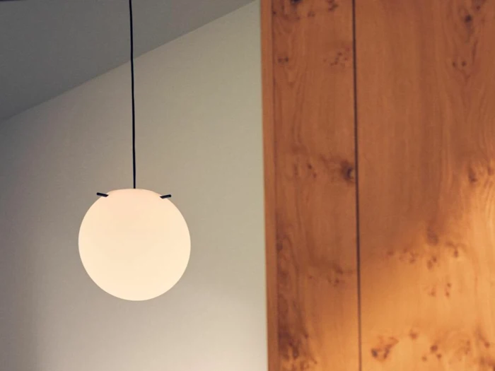 En rund, hvid pendel hænger fra loftet ved siden af en trævæg eller et træskab i en moderne, minimalistisk indretning.