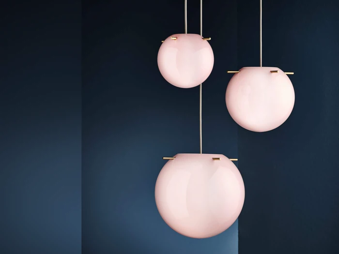 Tre moderne lyserøde glaspendler med gulddetaljer hænger i forskellige højder mod en mørkeblå væg og skaber et minimalistisk og elegant indretningslook.