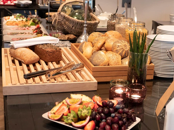 En morgenmadsbuffet med forskellige brød, rundstykker, krukker med korn, frisk frugt, tallerkener og stearinlys arrangeret på et bord.