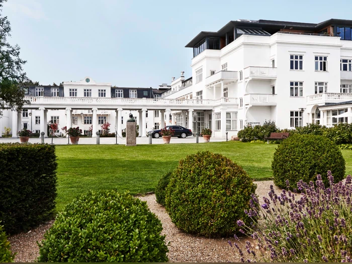 En stor hvid hotelbygning med balkoner og mange vinduer, omgivet af anlagte haver, grønne plæner, formklippede buske og blomstrende lavendler i forgrunden.
