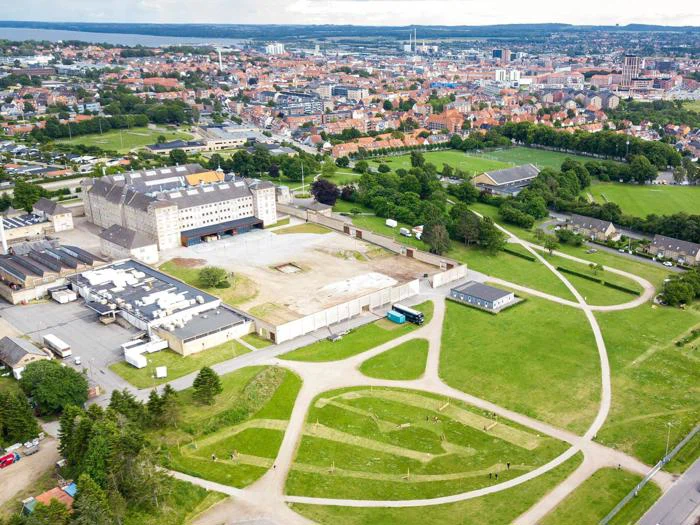 Luftfoto af en by med en stor historisk bygning, grønne parkområder, stier og omkringliggende boligkvarterer.