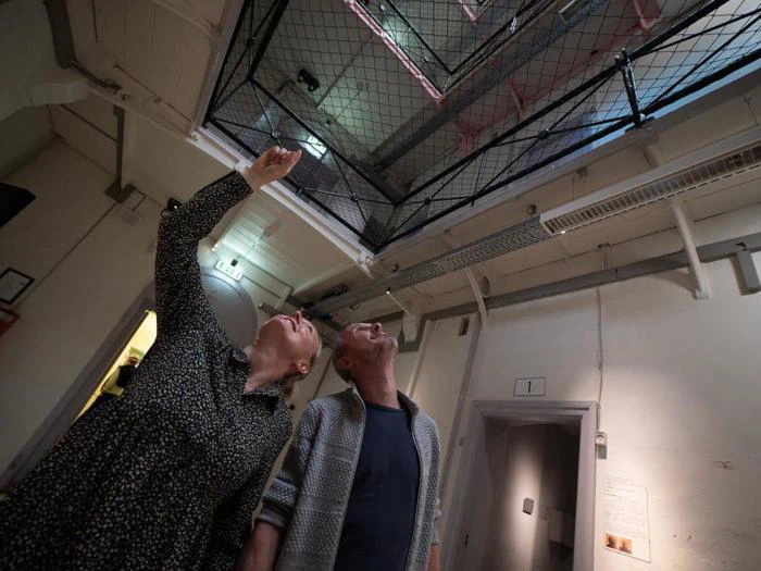 En kvinde og en mand står indendørs og kigger op og peger mod en netåbning i loftet, hvor der strømmer lys ned fra oven.