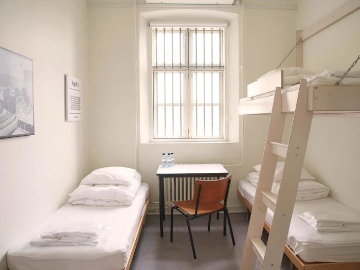 Et lille, lyst værelse med en køjeseng og en enkeltseng, et bord med to vandflasker, en træstol og et sprosset vindue, der lukker naturligt lys ind.