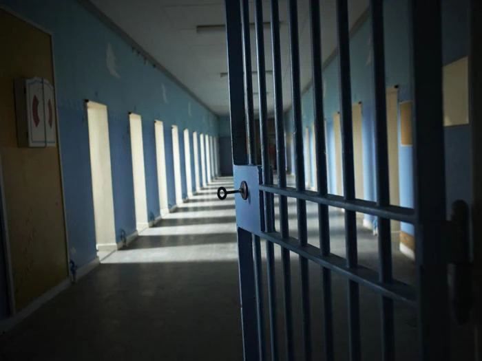 En svagt oplyst fængselsgang med en delvist åben metalport og sollys, der strømmer ind gennem sprossede vinduer langs den ene væg.