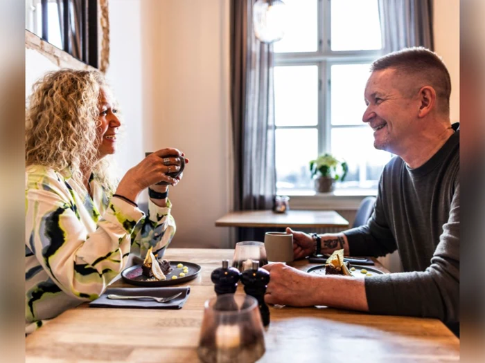 En kvinde og en mand sidder ved et bord i en lys café og smiler og taler, mens de nyder kaffe og dessert sammen.