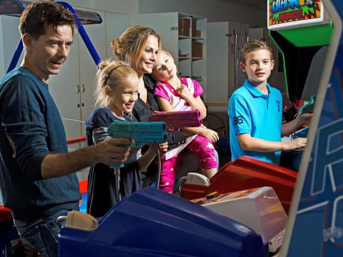 En smilende familie spiller arkadespil; to børn skyder med legetøjspistoler, mens en kvinde holder et yngre barn, og en dreng spiller ved en anden arkademaskine.