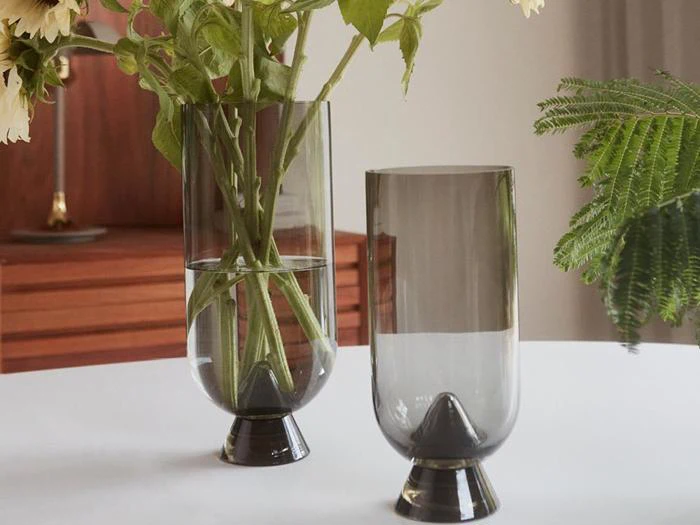 To moderne glasvaser, den ene med blomster og vand, står på en hvid flade ved siden af en grøn plante og træmøbler i baggrunden.