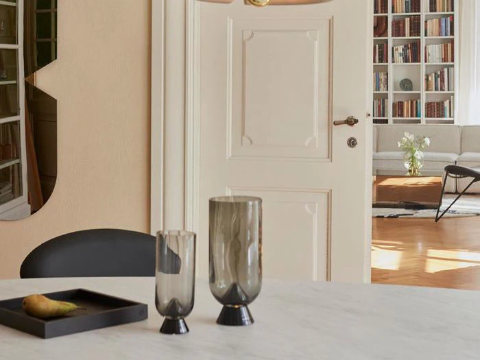 Et moderne spisebord med to grå glasvaser og en sort bakke med en pære, som står i et lyst rum med en åben dør og bogreoler i baggrunden.