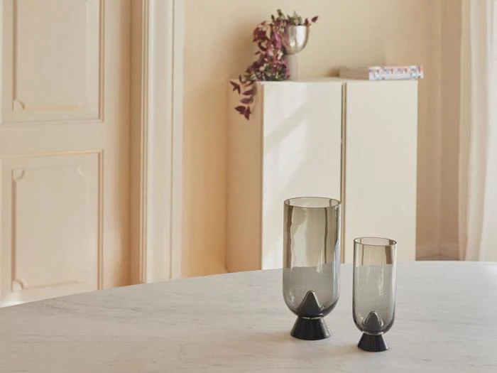 To moderne, røgfarvede glasvaser i forskellige højder står på et rundt, hvidt bord. I baggrunden står et hvidt skab med en plante i en sølvvase på toppen.