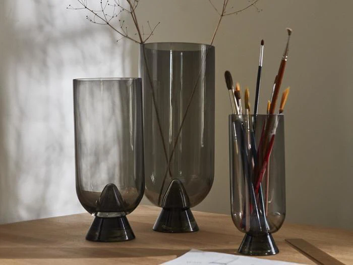 Tre røgfarvede glasvaser på en træplade; to indeholder tørrede grene, og en indeholder pensler. Blødt naturligt lys kaster skygger på væggen.
