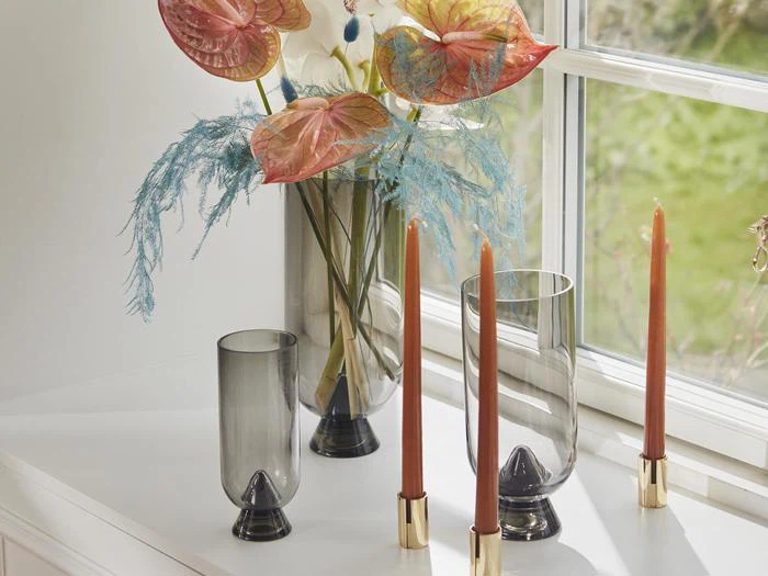 Røgfarvede glasvaser med rosa og hvide blomster og blåt løv står i vindueskarmen ved siden af tre brændte orange stearinlys i guldstager.