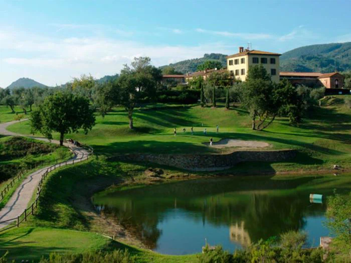 En naturskøn golfbane med frodigt grønt græs, en lille dam, spredte træer og en stor villa på en bakke i baggrunden under en klar blå himmel.