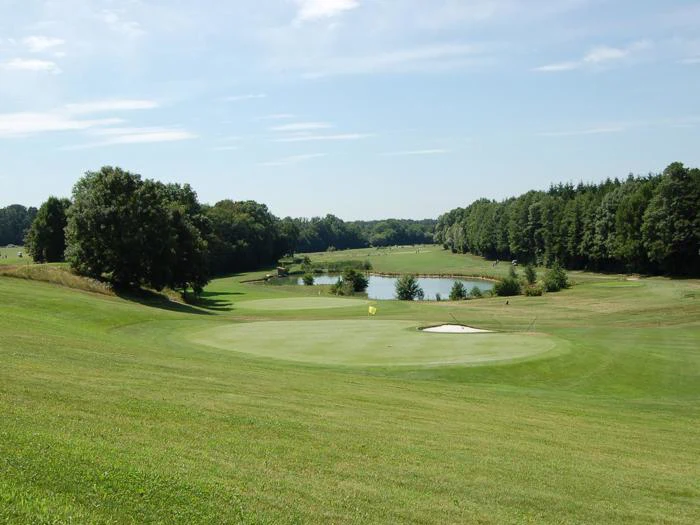 En grøn golfbane med en sandbunker, omgivet af træer og en dam i det fjerne under en delvist overskyet himmel.