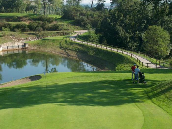 To personer står på en golfbane i nærheden af en golfbag, ved siden af en lille dam og en snoet sti med trægelænder. Træer og græs omgiver scenen.