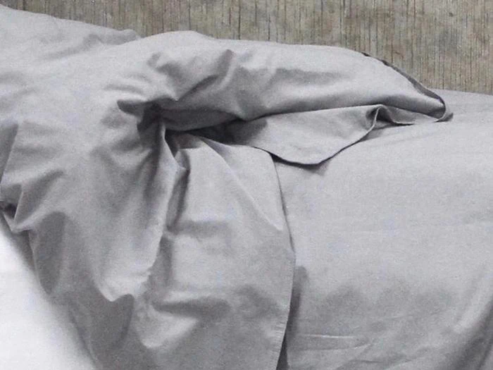 En pænt redt seng med lysegrå lagner og et tæppe, der er lidt krøllet i toppen, mod en struktureret grå væg.