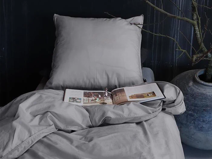 En seng med grå lagner og pude; et åbent magasin og briller ligger på betrækket, og der står en stor potteplante ved siden af sengen.
